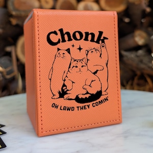 Peut inclure: Une boîte de rangement de cartes orange avec le mot "Chonk" au-dessus d'un dessin de trois chats de dessin animé. La phrase "OH LAWD THEY COMIN'" est imprimée en dessous. La boîte est sur une surface blanche.