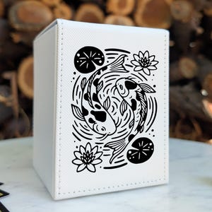 May include: A white rectangular deck box featuring a black graphic illustration of two koi fish swimming in a circular pattern, surrounded by water ripples, lily pads, and lotus flowers. The box has visible stitching along the edges.