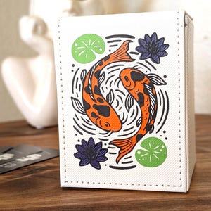 May include: A white rectangular deck box featuring a graphic illustration of two orange and black koi fish swimming in a circle. The design includes black line work representing water ripples, two green lily pads, and two dark purple lotus flowers.