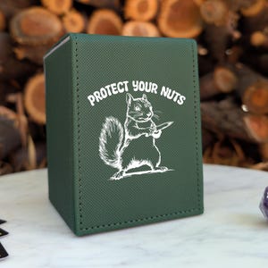 May include: A dark green rectangular deck box with a textured finish. The front features a white graphic of a squirrel holding a knife, accompanied by the text "PROTECT YOUR NUTS" arched above the squirrel in a bold, white, sans-serif font.