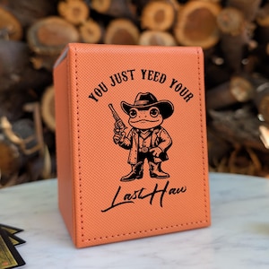 May include: An orange card storage box with the text "You Just Yeed Your Last Haw" and a cartoon frog wearing a cowboy hat and holding a gun. The box is sitting on a white surface with a few cards and a die.
