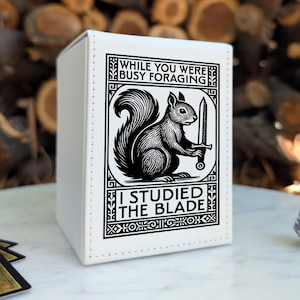 May include: A white rectangular deck box featuring a black graphic print of a squirrel holding a sword. The text reads: WHILE YOU WERE BUSY FORAGING I STUDIED THE BLADE. The box has visible stitching along the edges and a smooth, matte finish.