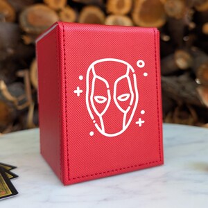 May include: A rectangular red deck box with a textured surface featuring a white line art graphic of a masked character face on the front panel. The box has stitched edges and is positioned on a white surface with game cards and a die nearby.