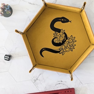 May include: A yellow hexagonal dice tray with a black snake and floral design. The tray has a suede-like texture and is made of leather.