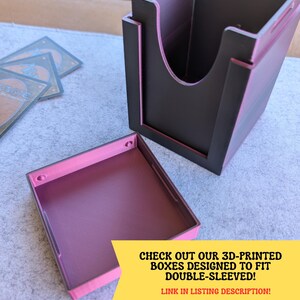 May include: A 3D-printed card storage box with a removable lid. The box is primarily black with pink accents and interior. The text "CHECK OUT OUR 3D-PRINTED BOXES DESIGNED TO FIT DOUBLE-SLEEVED! LINK IN LISTING DESCRIPTION!" is visible.
