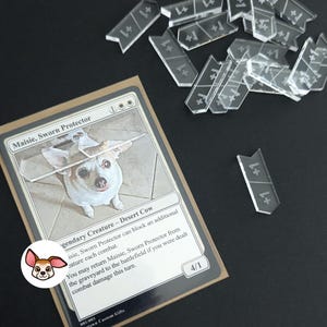 May include: A Magic: The Gathering card featuring a dog named Maisie, Sworn Protector, a legendary creature with the text "Desert Cow". The card is a 4/1 creature with the ability to block an additional creature each combat and return itself to the battlefield from the graveyard if the player was dealt combat damage this turn.