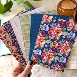 May include: A set of rectangular bookmarks featuring various patterns including a dark blue background with colorful roses, a blue textured design, a green plaid pattern, a light cream lace-like print, a dark purple floral print, and a tan floral design.