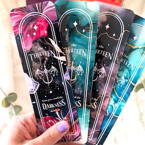 May include: A hand holds a collection of colorful bookmarks. Each bookmark features the text "the THIRTEEN" and "from now until the DARKNESS claims us." The bookmarks have a dark background with floral and abstract designs in shades of pink, teal, and blue.
