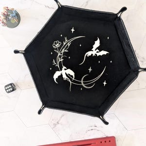 May include: Black hexagonal dice tray with a moon and two dragons design. The tray is made of felt and has a silver glitter finish.