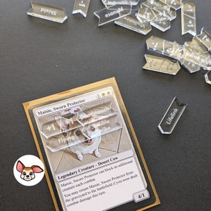 May include: A Magic: The Gathering card featuring a white dog with brown spots. The card is titled "Maisie, Sworn Protector" and is a Legendary Creature - Desert Cow. The card text reads: "Maisie, Sworn Protector can block an additional creature each combat. You may return Maisie, Sworn Protector from the graveyard to the battlefield if you were dealt combat damage this turn."