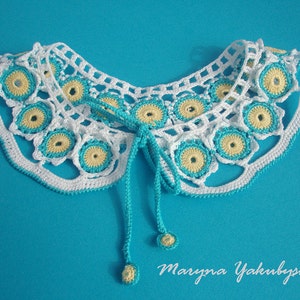 May include: White crocheted collar with blue and yellow accents. The collar has a tie closure and is on a blue background.