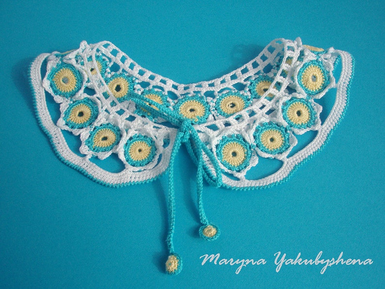 May include: White crocheted collar with blue and yellow accents. The collar has a tie closure and is on a blue background.