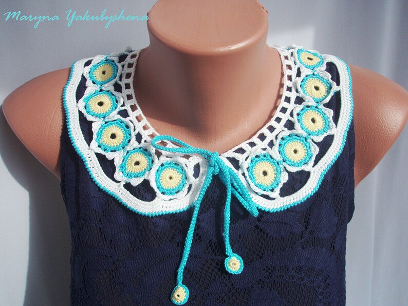 May include: A white crocheted collar with yellow and turquoise accents. The collar has a tie in the front and is worn on a blue sleeveless top.