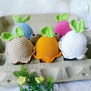 May include: Five crocheted Easter eggs in a cardboard egg carton. The eggs are decorated with green leaves and are in the colours blue, beige, white, pink, and yellow.