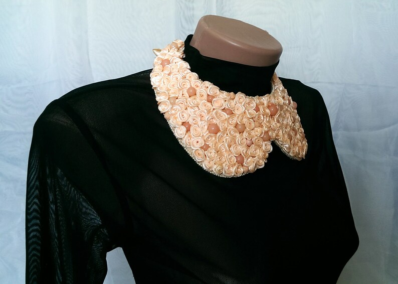 May include: A black sheer blouse with a large, peach-colored fabric flower collar. The collar is made of many small fabric roses and beads.