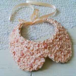May include: A peach-colored fabric collar necklace with a satin ribbon tie. The collar is made of many small fabric rosettes and has a scalloped edge.