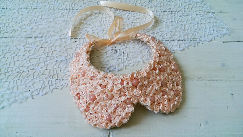 May include: A peach-colored fabric collar necklace with a satin ribbon tie. The collar is made of many small fabric rosettes and has a scalloped edge.