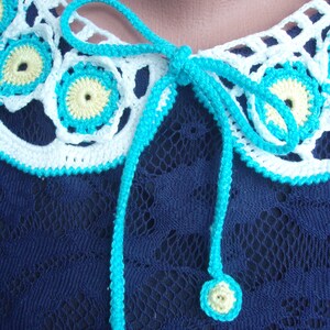 May include: A white crocheted collar with turquoise trim and yellow centers. The collar has a turquoise ribbon tied in a bow at the back.