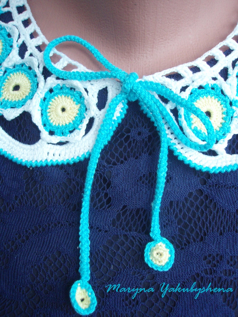 May include: A white crocheted collar with turquoise trim and yellow centers. The collar has a turquoise ribbon tied in a bow at the back.