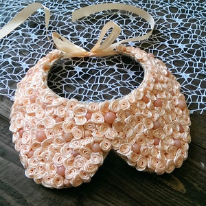 May include: A peach-colored fabric collar necklace with a satin ribbon tie. The collar is made of many small fabric rosettes and has a few pearl-like beads.