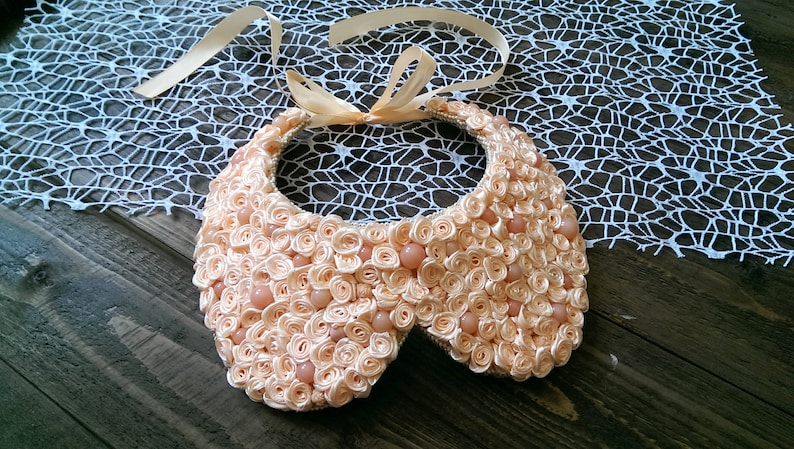 May include: A peach-colored fabric collar necklace with a satin ribbon tie. The collar is made of many small fabric rosettes and has a few pearl-like beads.