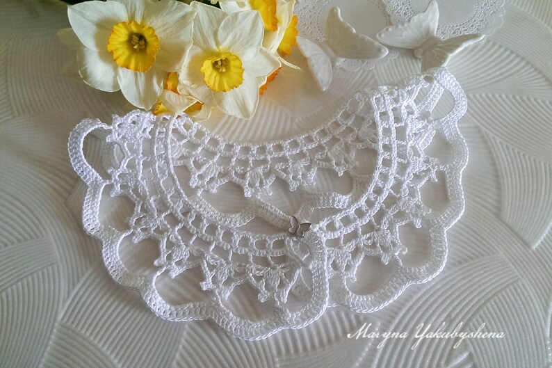 Puede incluir: Un cuello blanco de ganchillo con un borde festoneado y un delicado cierre de mariposa. El cuello se muestra con narcisos amarillos y blancos y decoraciones de mariposas blancas. Un accesorio hecho a mano.