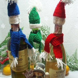 Puede incluir: Tres botellas de vino decoradas con gorros y bufandas de punto en rojo, verde y azul. Las botellas están rodeadas de una figura de muñeco de nieve, una rama de vegetación y pequeñas naranjas.