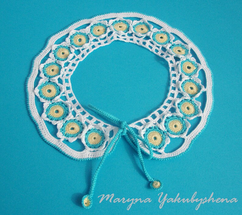 May include: White crocheted collar with blue and yellow accents. The collar has a tie closure and is decorated with a repeating pattern of small circles.