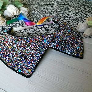 Peut inclure: Un collier en perles color&eacute;es avec un dos en tissu noir. Le collier est compos&eacute; de petites perles rondes de diff&eacute;rentes couleurs, notamment rouge, bleu, vert et blanc. Le collier est pos&eacute; sur une surface en bois blanc.