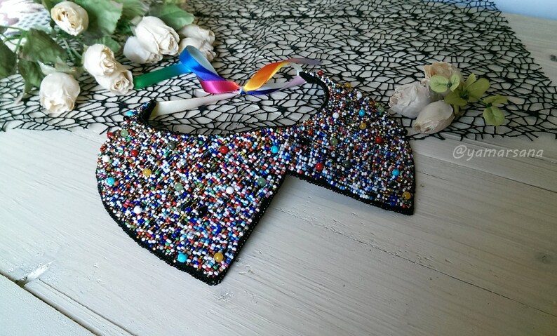 Peut inclure: Un collier en perles color&eacute;es avec un dos en tissu noir. Le collier est compos&eacute; de petites perles rondes de diff&eacute;rentes couleurs, notamment rouge, bleu, vert et blanc. Le collier est pos&eacute; sur une surface en bois blanc.