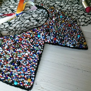 Peut inclure: Un collier de perles color&eacute; avec un dos en tissu noir. Le collier est compos&eacute; de petites perles multicolores, notamment bleues, vertes, rouges, blanches et dor&eacute;es. Le collier est pos&eacute; sur une surface en bois blanc.