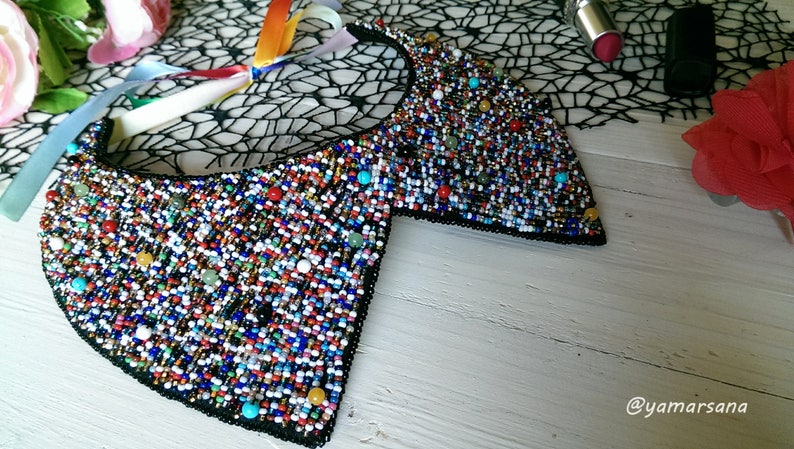 Peut inclure: Un collier de perles color&eacute; avec un dos en tissu noir. Le collier est compos&eacute; de petites perles multicolores, notamment bleues, vertes, rouges, blanches et dor&eacute;es. Le collier est pos&eacute; sur une surface en bois blanc.
