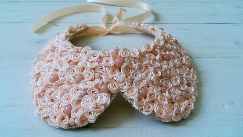 May include: A delicate peach-colored collar necklace made of fabric rosettes and pearl-like beads.