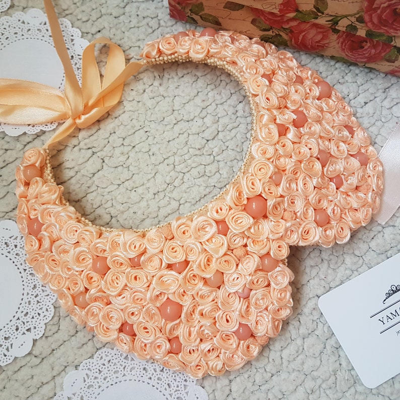 May include: Peach-colored fabric collar necklace with a large, ruffled design. The necklace is made of many small fabric rosettes and is decorated with small, round, peach-colored beads.