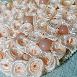 May include: A heart-shaped decoration made of peach-colored satin ribbon, with small pearl-like beads around the edge. The ribbon is arranged in a series of overlapping rosettes, creating a textured and delicate design.