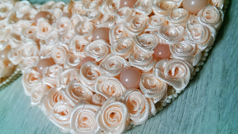 May include: A heart-shaped decoration made of peach-colored satin ribbon, with small pearl-like beads around the edge. The ribbon is arranged in a series of overlapping rosettes, creating a textured and delicate design.