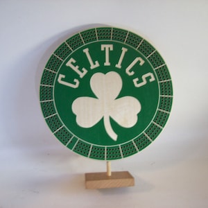 May include: A wooden cribbage board featuring the Boston Celtics logo. The design includes a green circle with the team name and a white shamrock. The board is supported by a wooden base and stand.