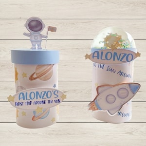 May include: Two blue and white cylindrical containers with space-themed designs. One container features an astronaut holding a flag with the name "Alonzo" on it. The other container has a rocket ship and the text "Alonzo's First Trip Around The Sun".
