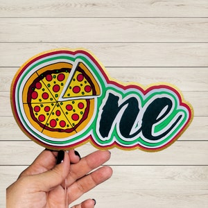 Puede incluir: Un adorno de pastel de papel colorido con la palabra "one" en letras negras y verdes y una rebanada de pizza con pepperoni rojo y pimientos verdes sobre una corteza amarilla.