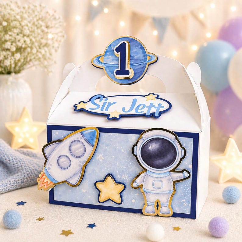 May include: A white gift box with a blue and gold space theme. The box features a rocket, astronaut, and star decorations. The text "Sir Jett" is displayed on the front, with a number "1" on top. Balloons and decorative stars are in the background.
