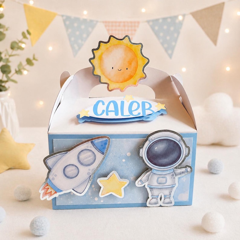 May include: A white box with a blue and white space theme. The box features a rocket, astronaut, and star stickers. The top has a sun and the name "CALEB" in blue. Decorative bunting and lights are in the background.
