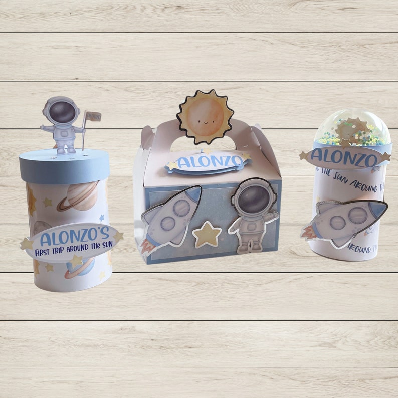 May include: Three space-themed party favor boxes. One is a cylinder with an astronaut on top and the text "ALONZO'S FIRST TRIP AROUND THE SUN". Another is a box with a sun and astronaut. The third is a cup with a rocket.
