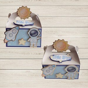 May include: Two white favor boxes with blue panels featuring a rocket, astronaut, and star. Each box has a sun-shaped topper and a label that reads "Zayne Apollo". The boxes are decorated with space-themed cutouts.