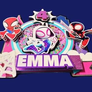 May include: A multi-layered cake topper featuring Spider-Man characters and the name "EMMA". The design includes a pink and white color scheme, a number "4", and a glittery purple banner. The background is a dark blue.