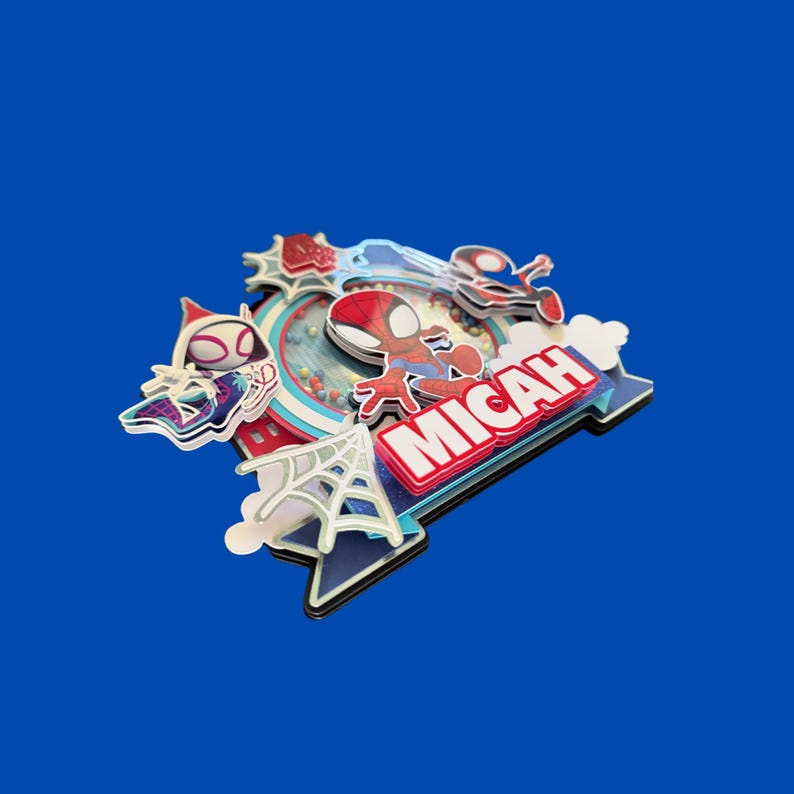 May include: A multi-layered cake topper featuring Spider-Man characters. The design includes a red and blue Spider-Man figure, a spider web, and the name "MICAH" in red and white. The topper has a blue and white color scheme with a blue background.