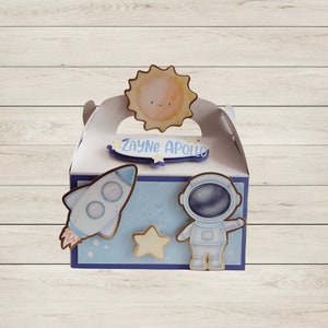 May include: A white gift box with a blue and white space theme. The box features a rocket, astronaut, star, and sun cutouts. The name "Zayne Apollo" is written on a blue banner. The box has a handle and is decorated with a space theme.
