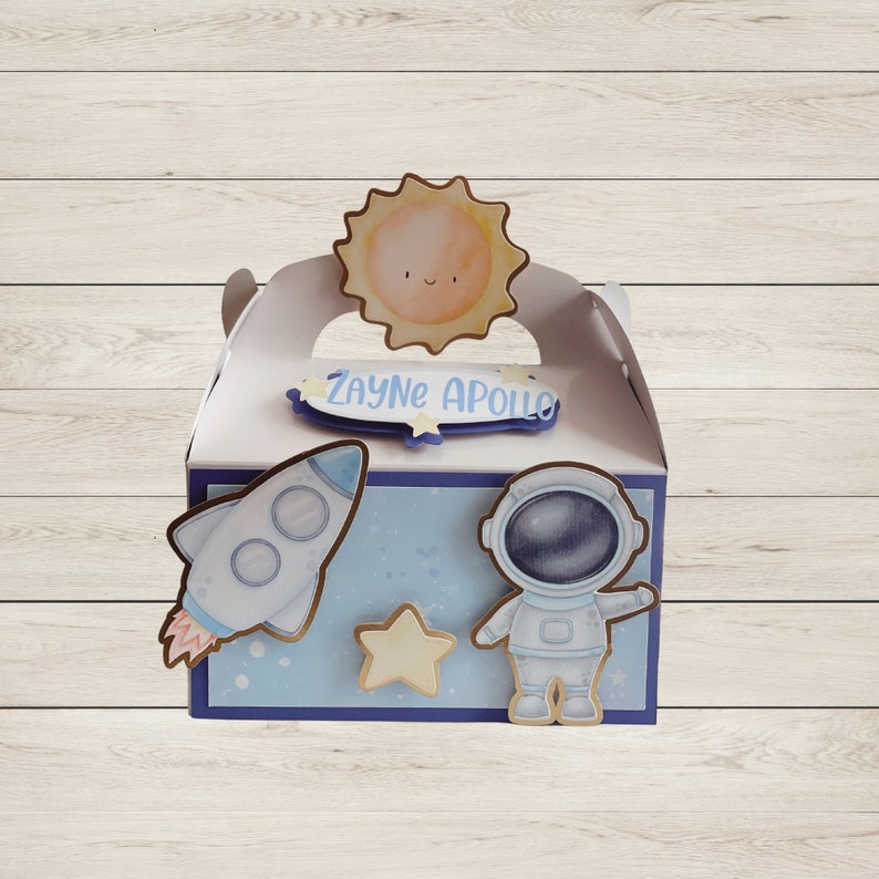 May include: A white gift box with a blue and white space theme. The box features a rocket, astronaut, star, and sun cutouts. The name "Zayne Apollo" is written on a blue banner. The box has a handle and is decorated with a space theme.