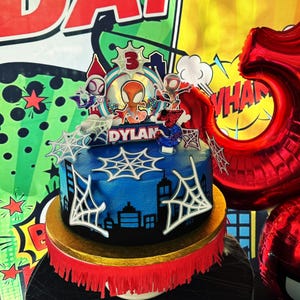 May include: A blue birthday cake decorated with a Spider-Man theme. The cake features white spiderwebs, a cityscape silhouette, and a topper with Spider-Man figures and the name "Dylan". The cake is on a gold and red base.