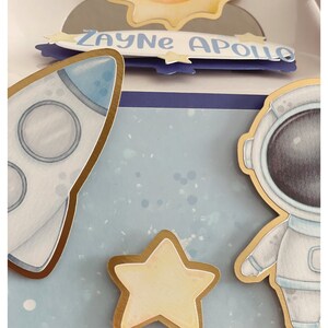 May include: A space-themed papercraft design featuring a rocket, astronaut, and star, all with gold trim. The name "Zayne Apollo" is written on a blue banner. The background is a light blue with white speckles.