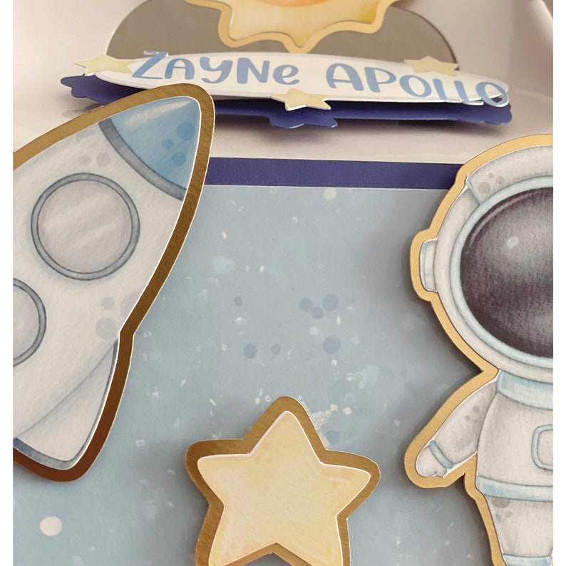 May include: A space-themed papercraft design featuring a rocket, astronaut, and star, all with gold trim. The name "Zayne Apollo" is written on a blue banner. The background is a light blue with white speckles.